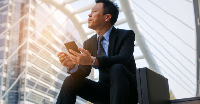 Asian man in a suit sitting on steps with a brief case and phone in hand looking up to the sky