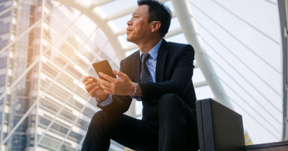 Asian man in a suit sitting on steps with a brief case and phone in hand looking up to the sky