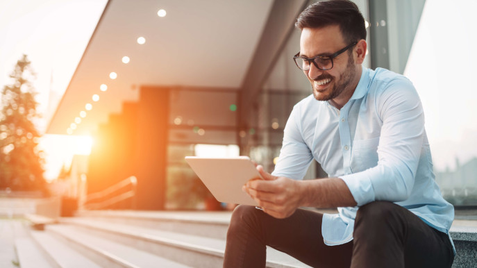 man smiling with glasses wearing smart clothes sitting outside on steps with tablet in hand