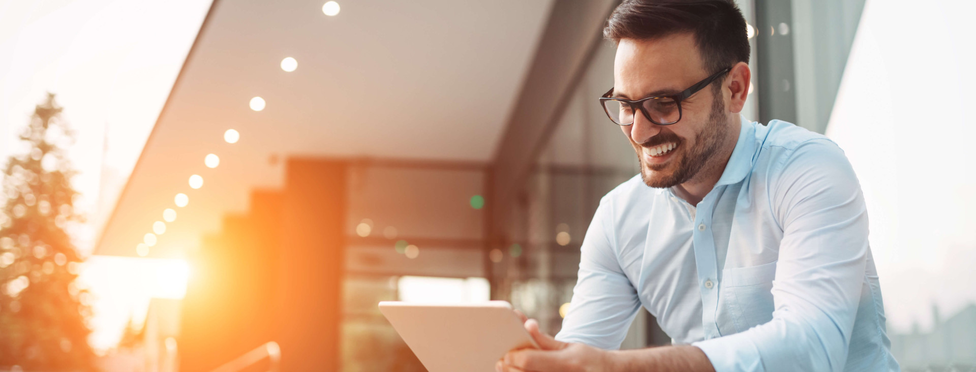 man smiling with glasses wearing smart clothes sitting outside on steps with tablet in hand