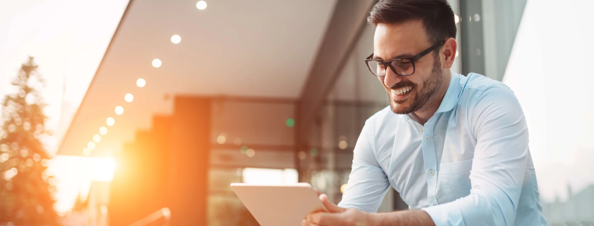 man smiling with glasses wearing smart clothes sitting outside on steps with tablet in hand