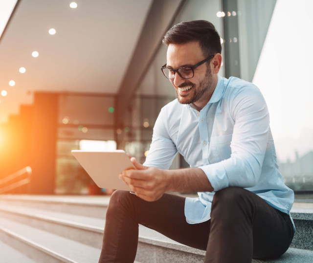 man smiling with glasses wearing smart clothes sitting outside on steps with tablet in hand