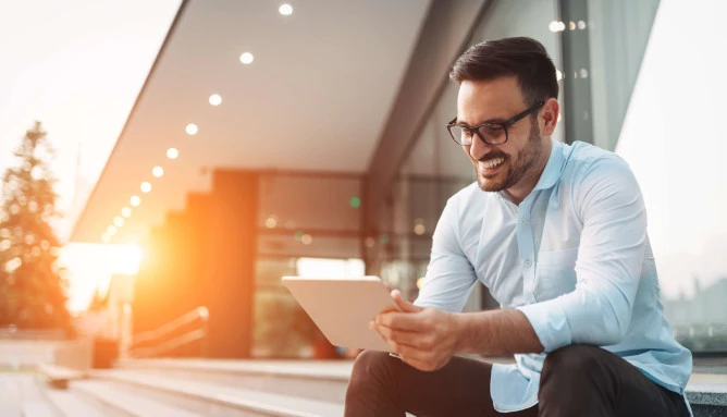 man smiling with glasses wearing smart clothes sitting outside on steps with tablet in hand