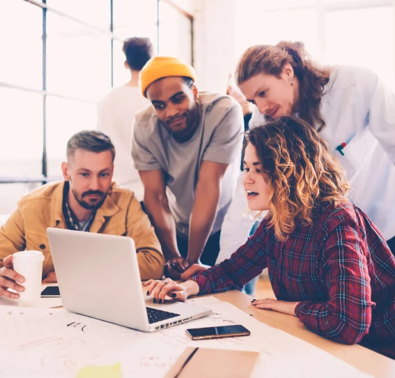 four people in causal clothing at a table in an office looking at a laptop