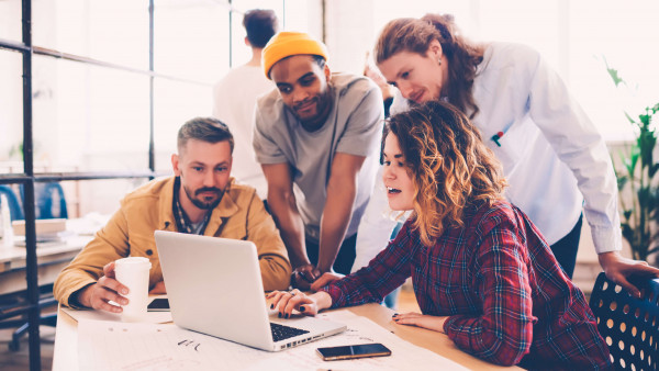 four people in causal clothing at a table in an office looking at a laptop
