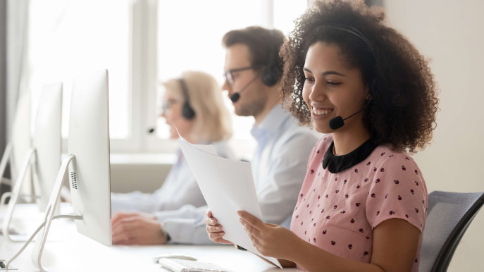woman at a desk in a call center with headset on holding documents with two people in background