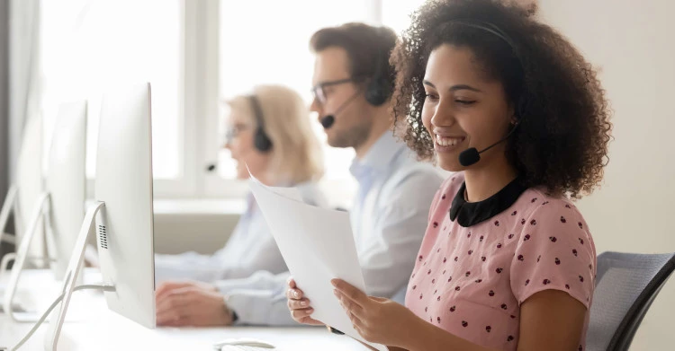 woman at a desk in a call center with headset on holding documents with two people in background