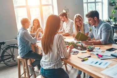 six people sitting in a communal area smiling and interacting with office material on the table
