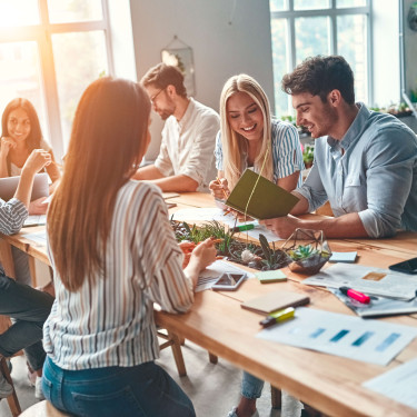 six people sitting in a communal area smiling and interacting with office material on the table