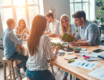 six people sitting in a communal area smiling and interacting with office material on the table