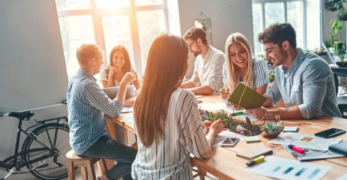 six people sitting in a communal area smiling and interacting with office material on the table