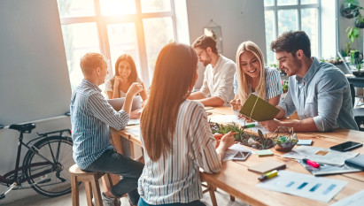 six people sitting in a communal area smiling and interacting with office material on the table