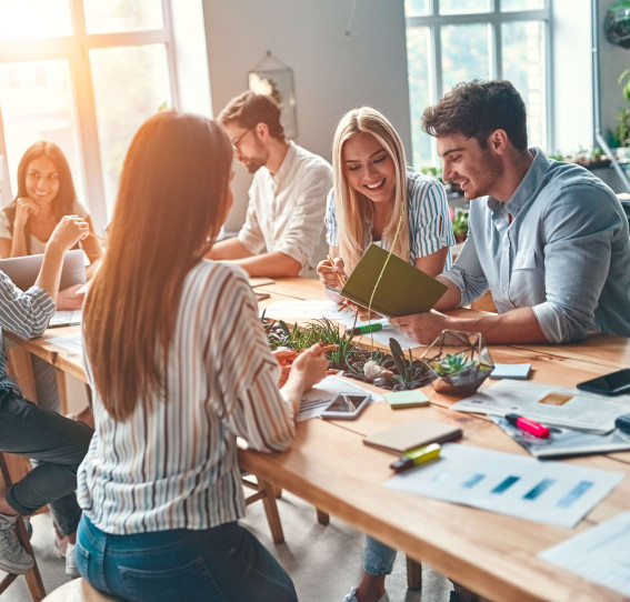 six people sitting in a communal area smiling and interacting with office material on the table