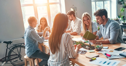 six people sitting in a communal area smiling and interacting with office material on the table