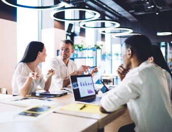 four people in smart clothing sitting in a office communal area with laptops and documents
