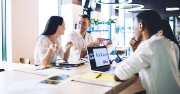 four people in smart clothing sitting in a office communal area with laptops and documents