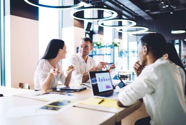 four people in smart clothing sitting in a office communal area with laptops and documents