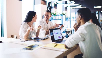 four people in smart clothing sitting in a office communal area with laptops and documents