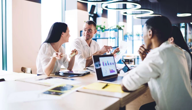 four people in smart clothing sitting in a office communal area with laptops and documents
