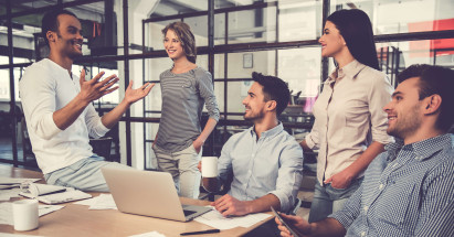 five people smiling gathered at a desk with a laptop and some paper