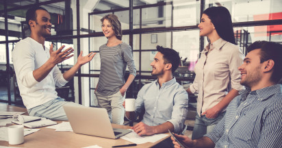 five people smiling gathered at a desk with a laptop and some paper