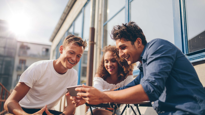 three people sitting outside smiling and looking at a mans mobile phone