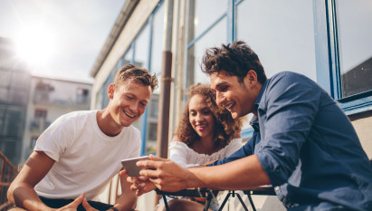 three people sitting outside smiling and looking at a mans mobile phone