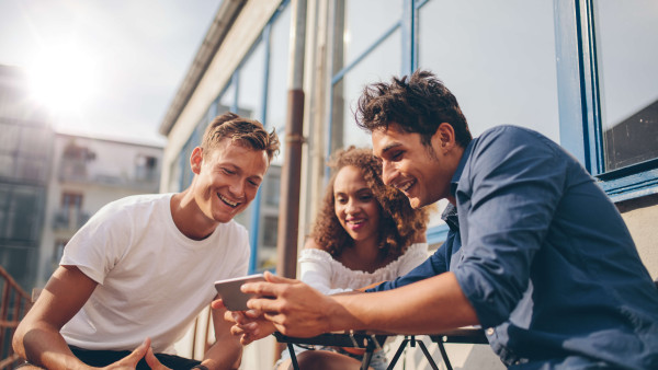 three people sitting outside smiling and looking at a mans mobile phone