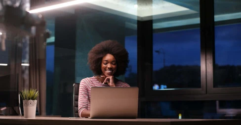 woman working in the evening smiling and sitting at a desk in an office with a laptop
