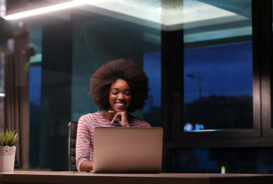 woman working in the evening smiling and sitting at a desk in an office with a laptop