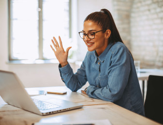 woman in smart casual clothing sitting at desk in front of laptop smiling and waving