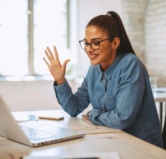 woman in smart casual clothing sitting at desk in front of laptop smiling and waving
