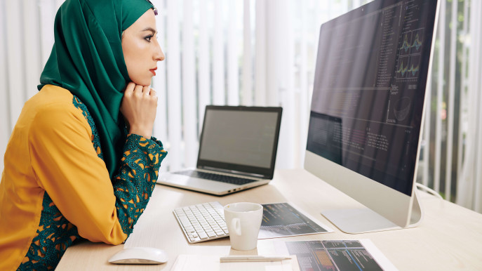 woman in hijab analysing data on apple mac with laptop and notes on the desk