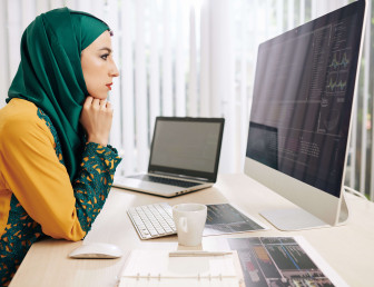 woman in hijab analysing data on apple mac with laptop and notes on the desk