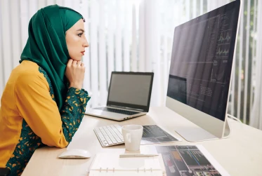 woman in hijab analysing data on apple mac with laptop and notes on the desk
