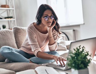 woman with glasses wearing casual clothes sitting with legs crossed on sofa looking at a laptop