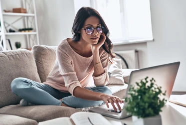 woman with glasses wearing casual clothes sitting with legs crossed on sofa looking at a laptop