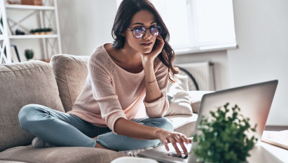 woman with glasses wearing casual clothes sitting with legs crossed on sofa looking at a laptop