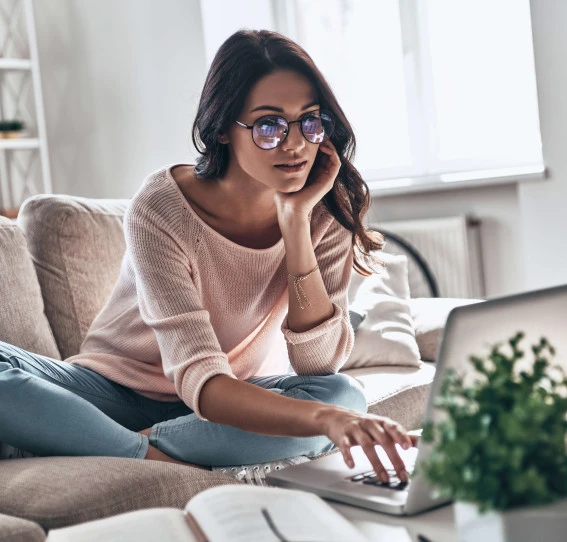 woman with glasses wearing casual clothes sitting with legs crossed on sofa looking at a laptop