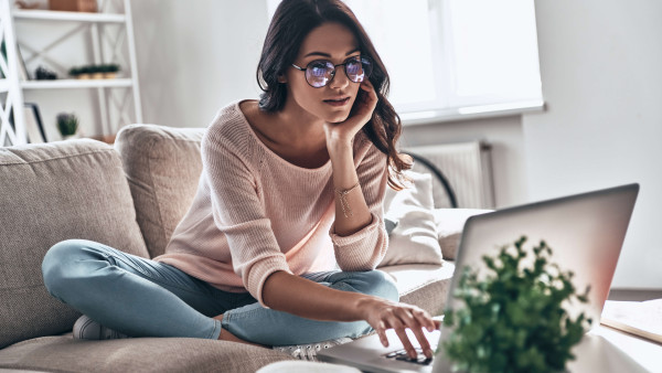 woman with glasses wearing casual clothes sitting with legs crossed on sofa looking at a laptop
