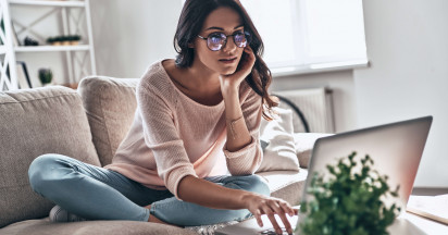 woman with glasses wearing casual clothes sitting with legs crossed on sofa looking at a laptop
