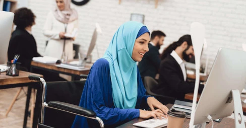 smiling woman in a wheelchair wearing a blue hijab working at a desk in an office