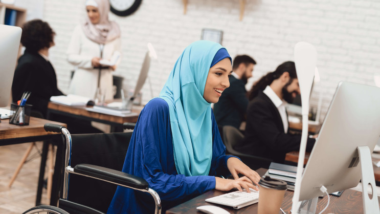 smiling woman in a wheelchair wearing a blue hijab working at a desk in an office