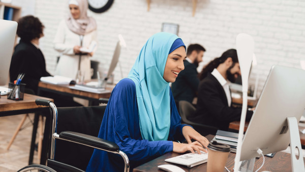 smiling woman in a wheelchair wearing a blue hijab working at a desk in an office