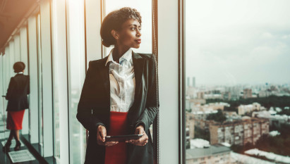 woman in suit looking out of office window holding tablet device