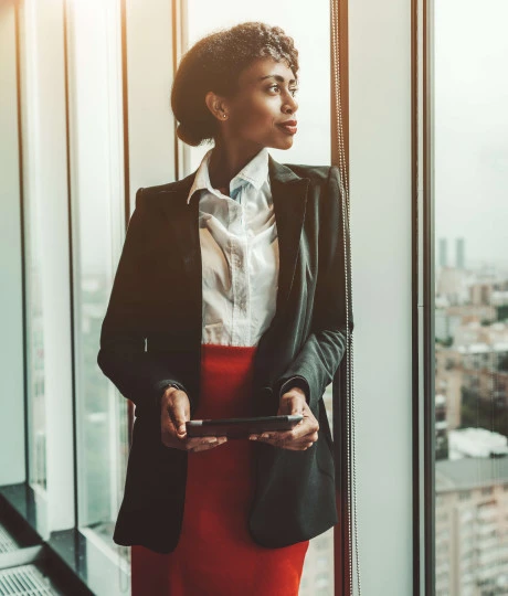 woman in suit looking out of office window holding tablet device