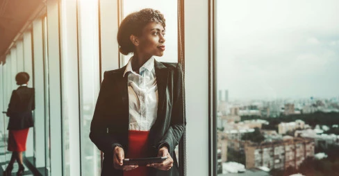 woman in suit looking out of office window holding tablet device