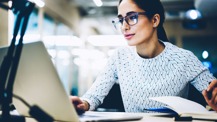 woman with glasses looking at laptop with pen in hand