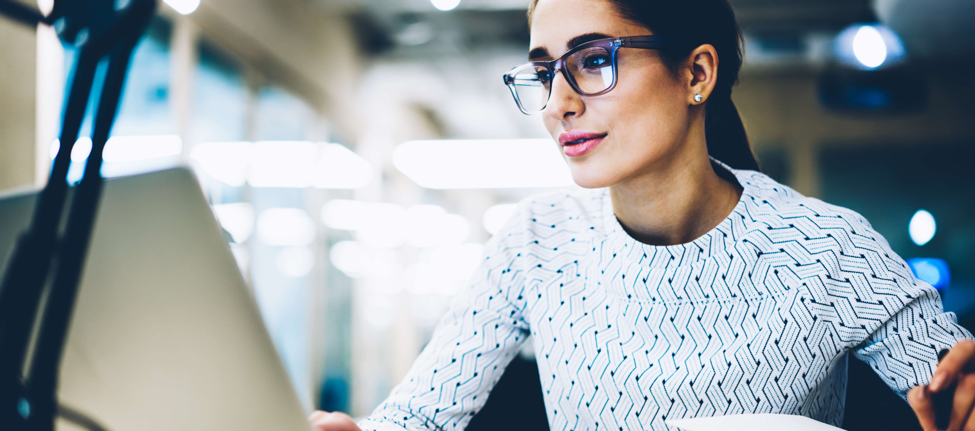 woman with glasses looking at laptop with pen in hand