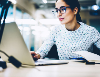 woman with glasses looking at laptop with pen in hand
